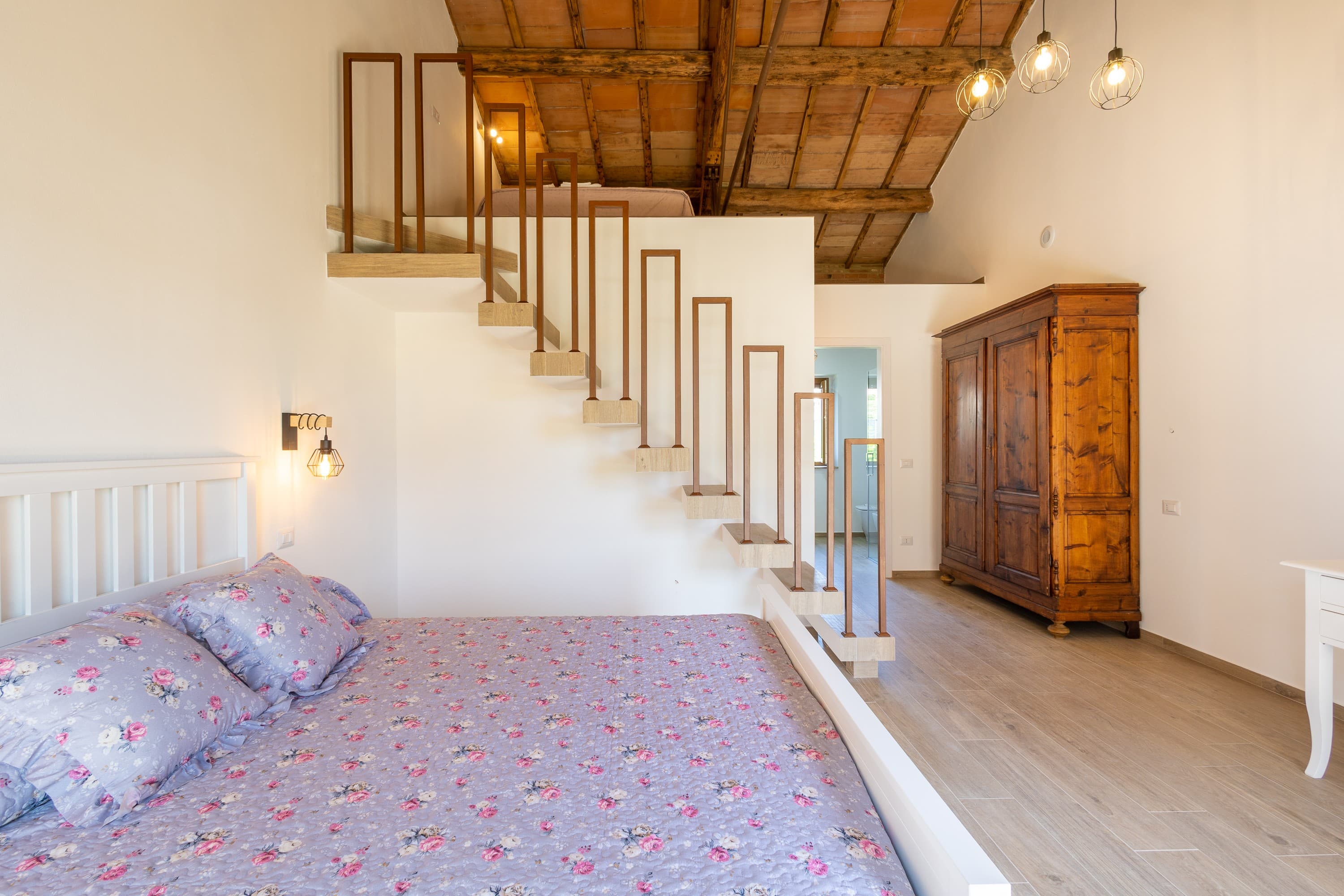 Spacious interior of a double bedroom featuring a high sloped ceiling with traditional rustic wooden beams and terracotta tiles. In the foreground, a double bed is dressed in a lilac floral print quilt and matching pillows. A modern suspended staircase with a minimalist geometric railing leads up to a mezzanine loft sleeping area. In the background, a large antique solid wood wardrobe sits against the wall near the bathroom entryway, complemented by a modern pendant light fixture with exposed filament bulbs. The room has light wood-effect tile flooring.