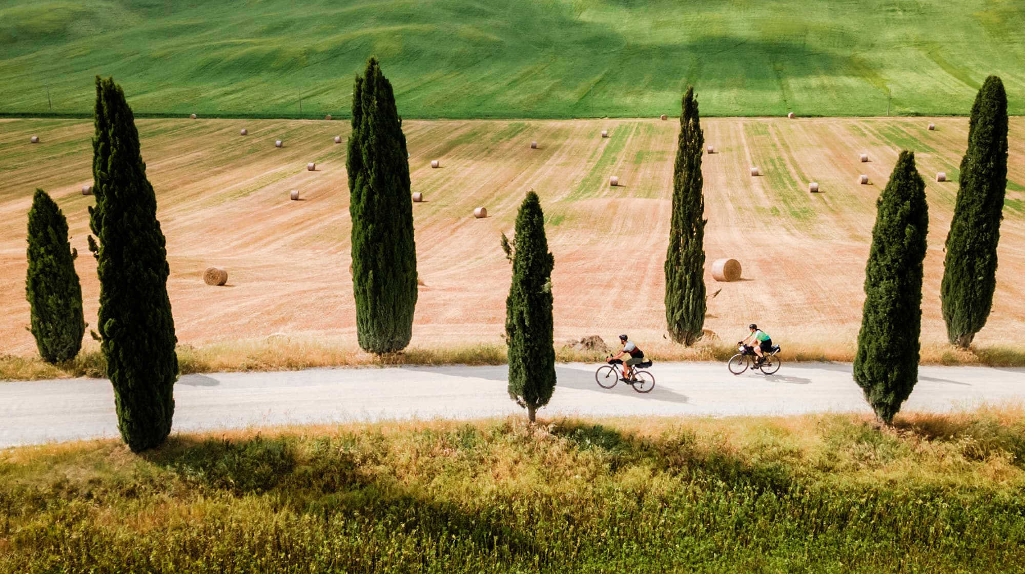 Deux participants au Tuscany Trail pédalent sur une route de campagne entre des rangées de cyprès.
