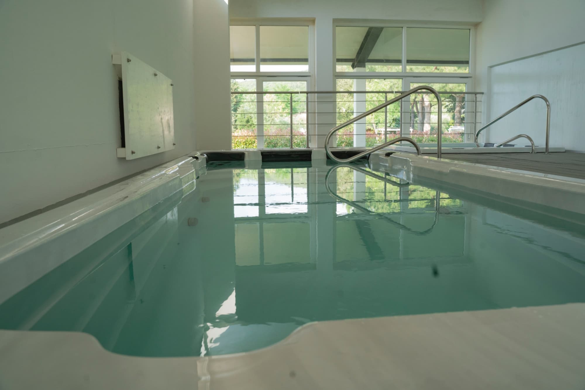 A low-angle view of a large rectangular indoor hot tub with crystal-clear water and white trim, set into a floor of dark wooden planks. In the background is a stainless steel handrail next to the access steps and, on a raised level, a second, smaller hot tub. Large floor-to-ceiling windows flood the space with light and offer views of the outdoor garden, creating a relaxing and bright spa-like atmosphere. 