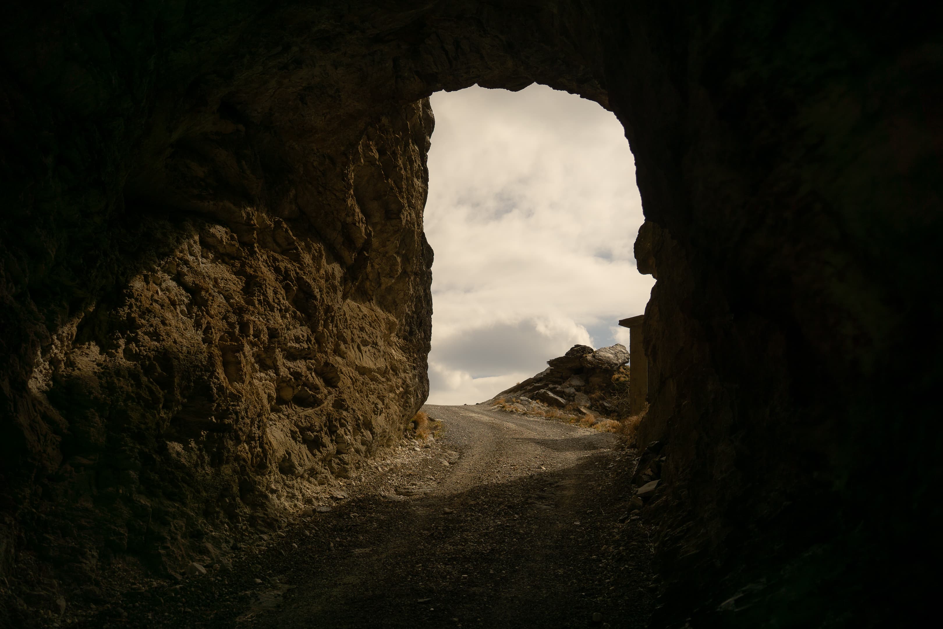 Galleria dug into the rock along the Marmifera del Corchia opens onto a white dirt mountain road, with rough stone walls on the sides, cloudy skies, and the rocky peaks of the Apuan Alps in the background