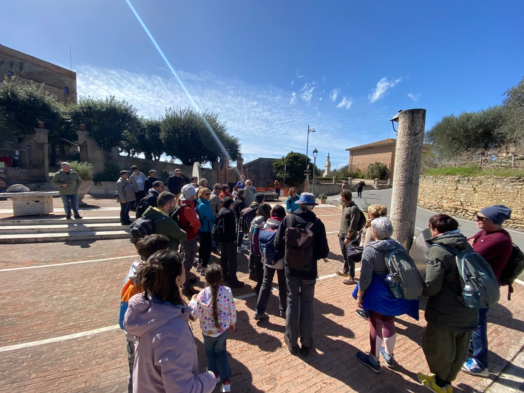 Gruppo di visitatori con zaini e bambini riuniti in una piazza in mattoni del borgo di San Giovanni d'Asso, in una giornata di sole autunnale, durante la visita guidata legata all'escursione del Treno Natura