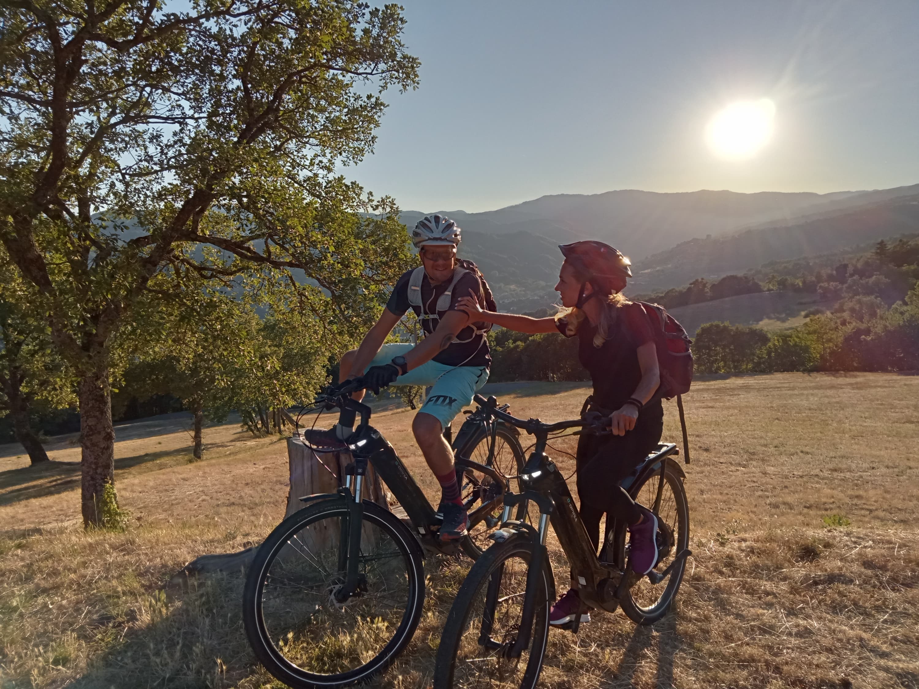 Couple of cyclists with helmets and backpacks aboard their e-bikes on an open hilly meadow, conversing at sunset with a panorama of Tuscan hills in the background and the sun low on the horizon