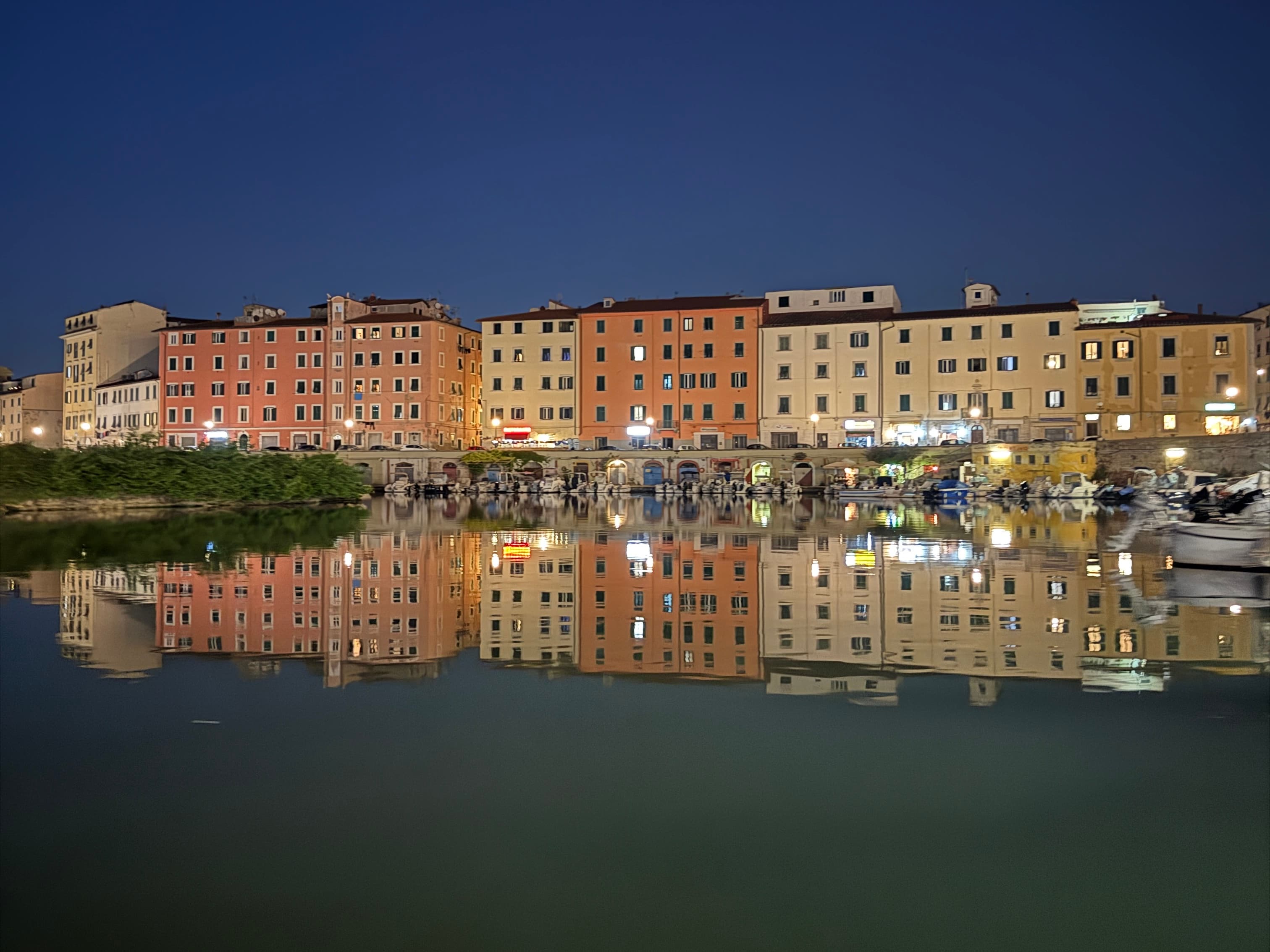 Livorno's waterfront at dusk with the silhouette of colourful multi-storey buildings reflected in the calm harbour water, boats moored on the right