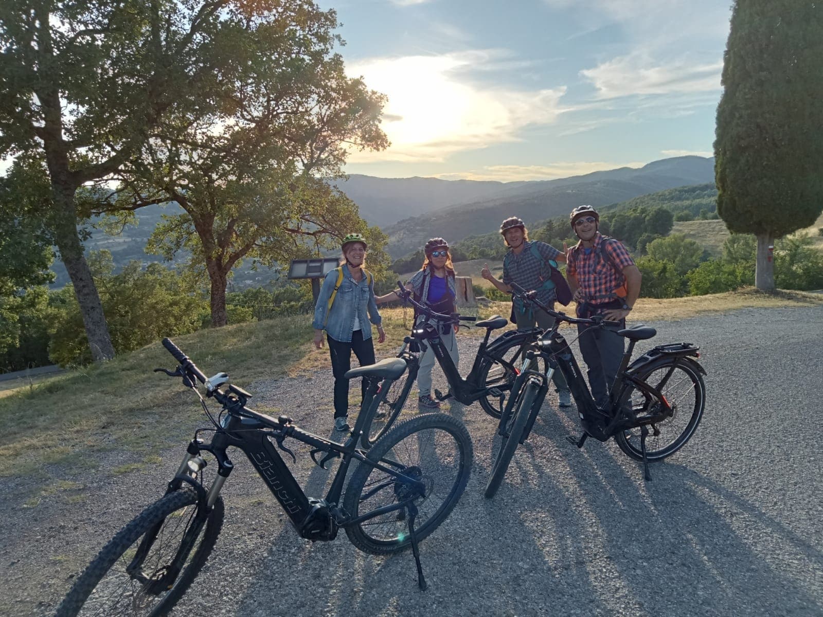 Group of four cyclists with helmets and Bianchi e-bikes stopped on a scenic road at sunset, with the wooded Casentino hills and mountains in the background