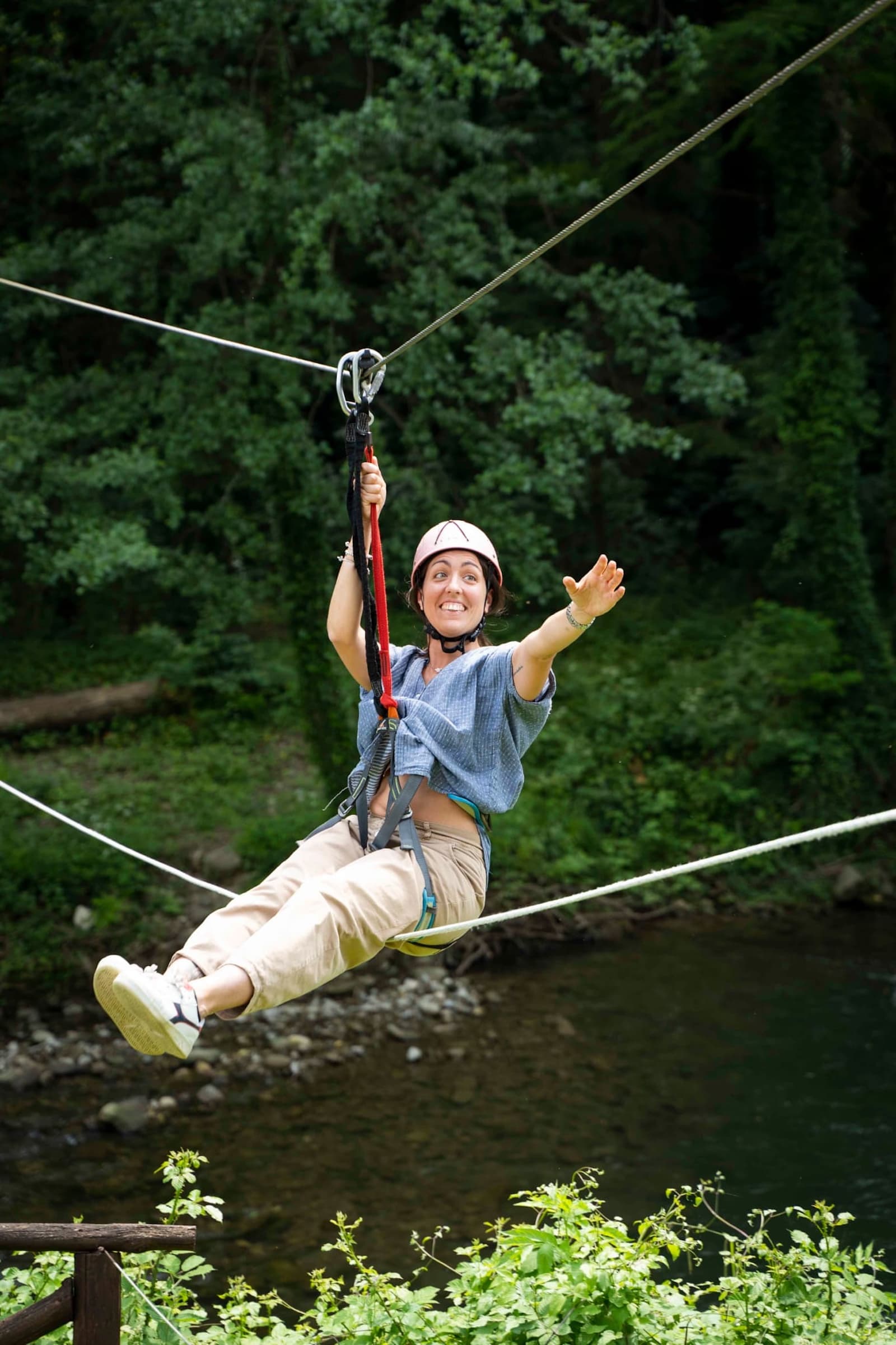 Ragazza sorridente con casco rosa e imbragatura che scivola su una teleferica del parco avventura, sospesa tra alberi verdi sopra il fiume Arno a Stia