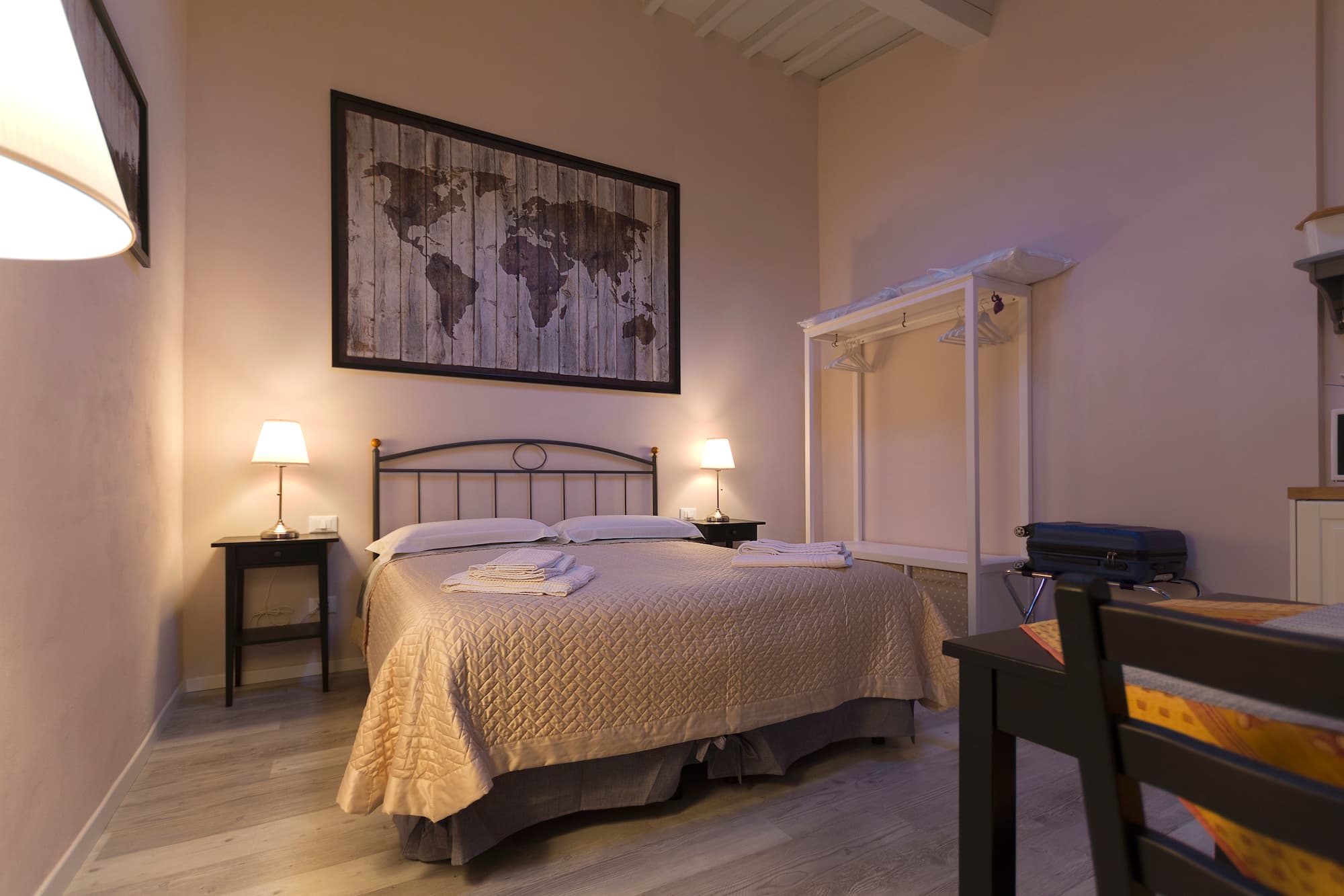 Interior view of a spacious and welcoming double bedroom with a blend of rustic and modern design. In the center a double bed features a dark wrought-iron headboard, a beige quilted bedspread, and neatly folded white towels for guests. On either side, black nightstands hold lighted table lamps emitting a warm glow. Hanging above the headboard is a large vintage-style world map printed on wooden planks. To the right, the room includes a white open wardrobe stand and a luggage rack holding a suitcase. The space is completed with light-colored parquet flooring and high ceilings with exposed whitewashed wooden beams.