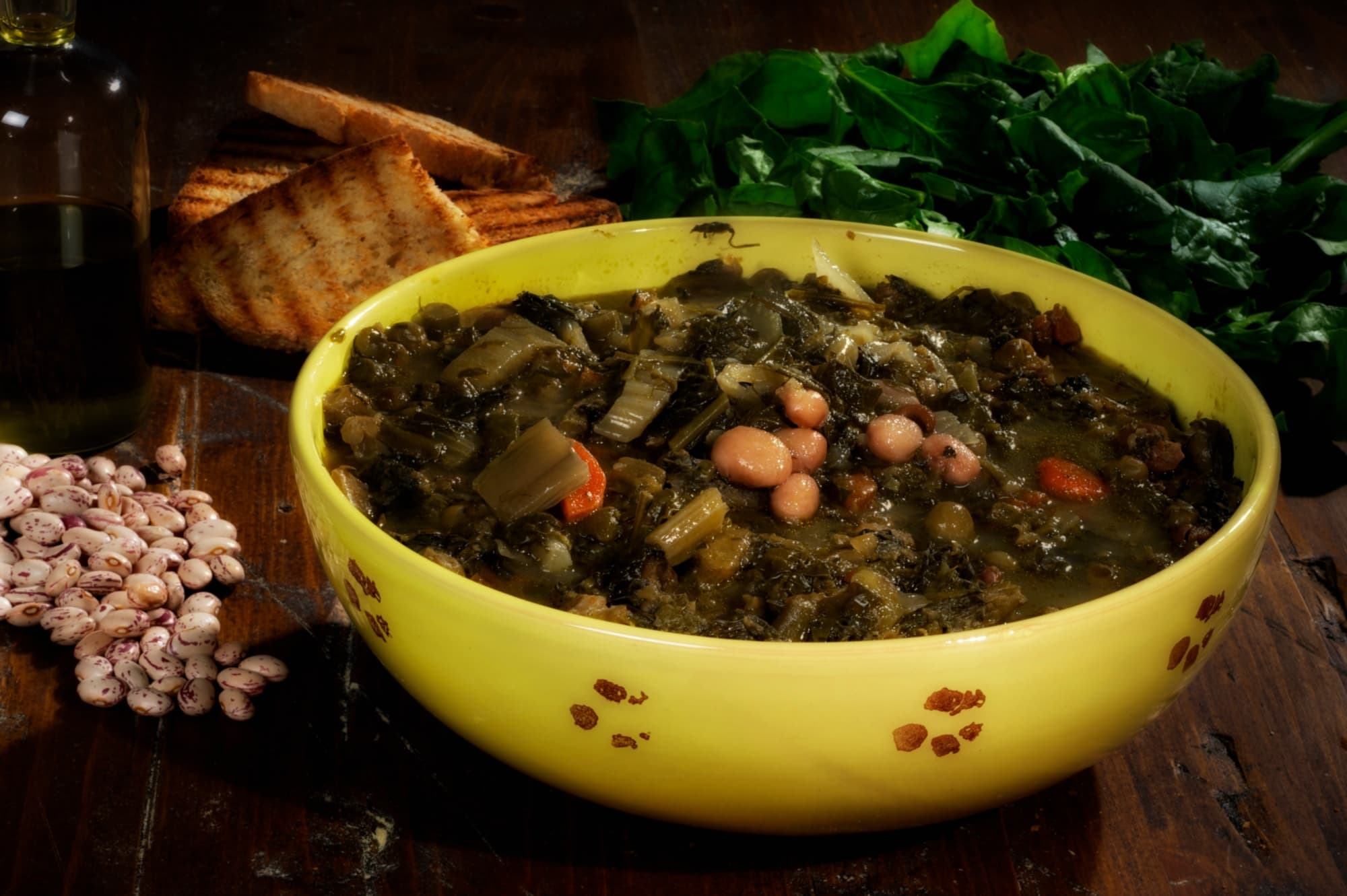 A ceramic plate containing traditional Garfagnana herb and bean soup. Around the plate: a few slices of toasted bread and a handful of dried beans