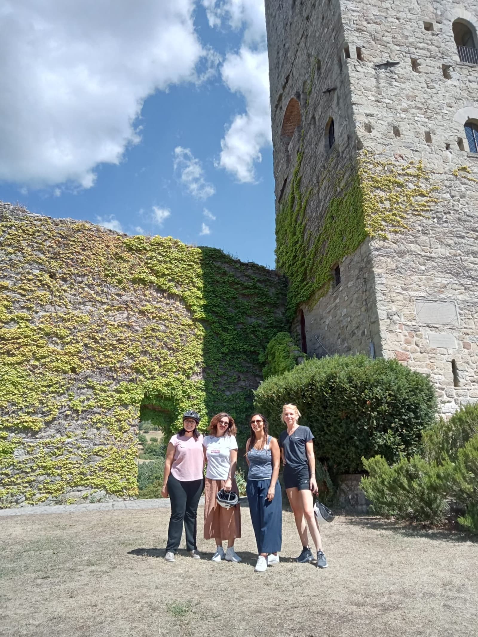 Four women in casual clothing, some holding bike helmets, pose smiling in front of the ivy-covered medieval stone tower of Porciano Castle during an e-bike excursion in Casentino.