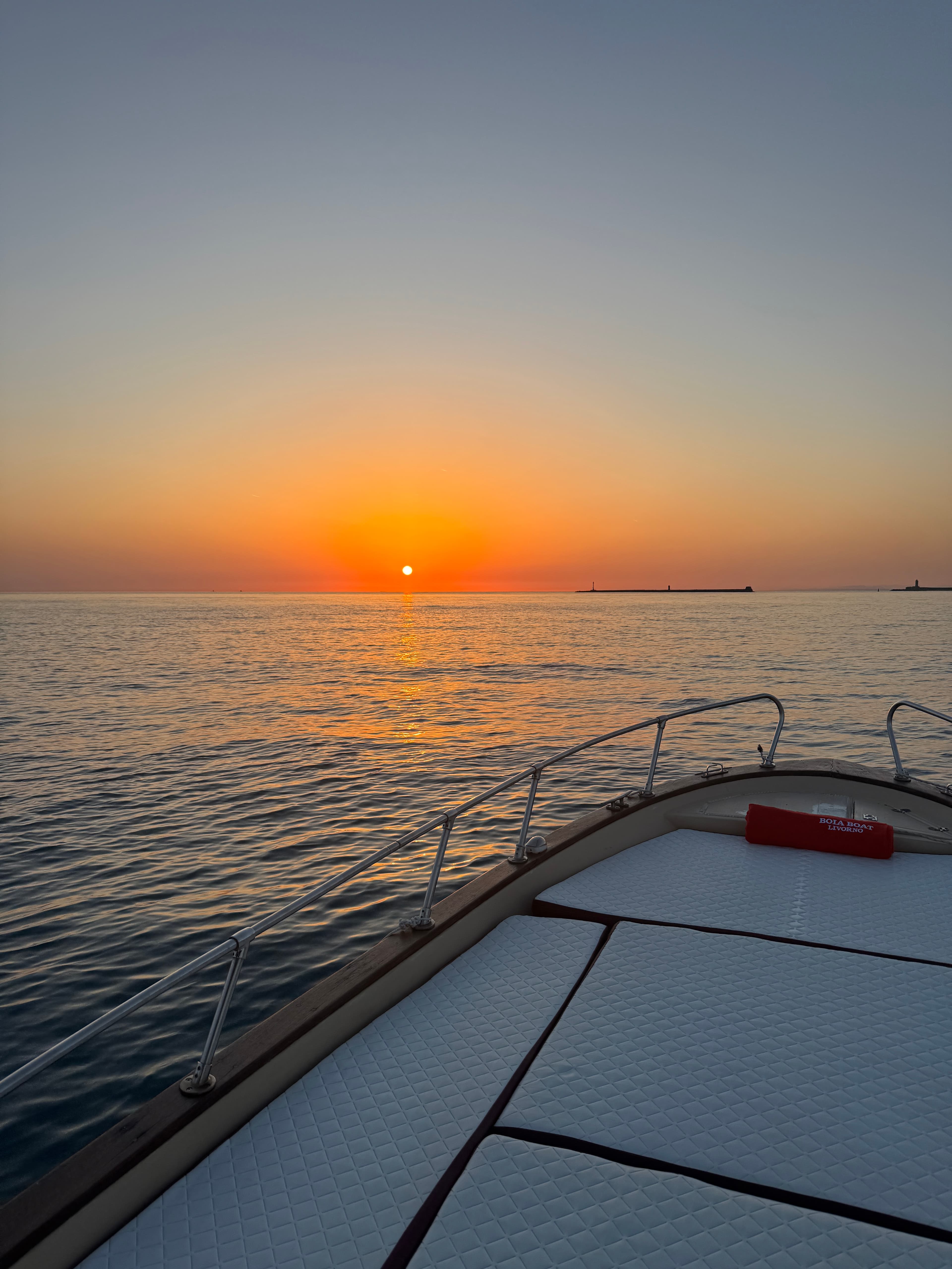 Bow of the Boia Boat Livorno with a red branded towel on the white sundeck, taken during a sunset over the sea with an orange sky and the outline of the port of Livorno on the horizon