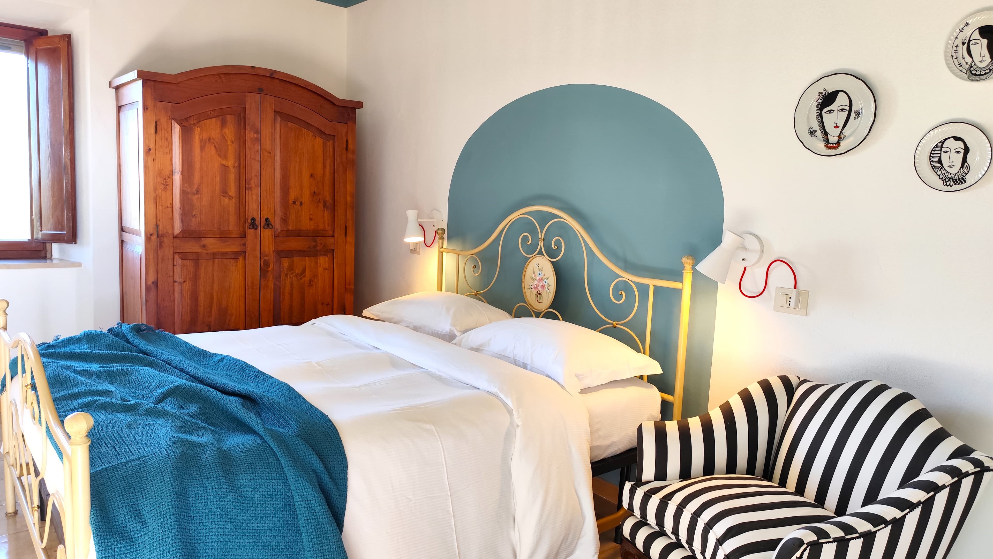 This image features a cozy bedroom that blends rustic and modern elements. The double bed has a cream-yellow wrought iron frame with an ornate floral medallion and a teal blanket draped over the white sheets. Behind the bed, a teal arch painted on the wall frames the headboard. Above the black and white striped upholstered armchair in the bottom right corner, there are three decorative black and white ceramic plates featuring stylized female faces. To the left, a large dark wood two-door wardrobe stands out next to an open window.