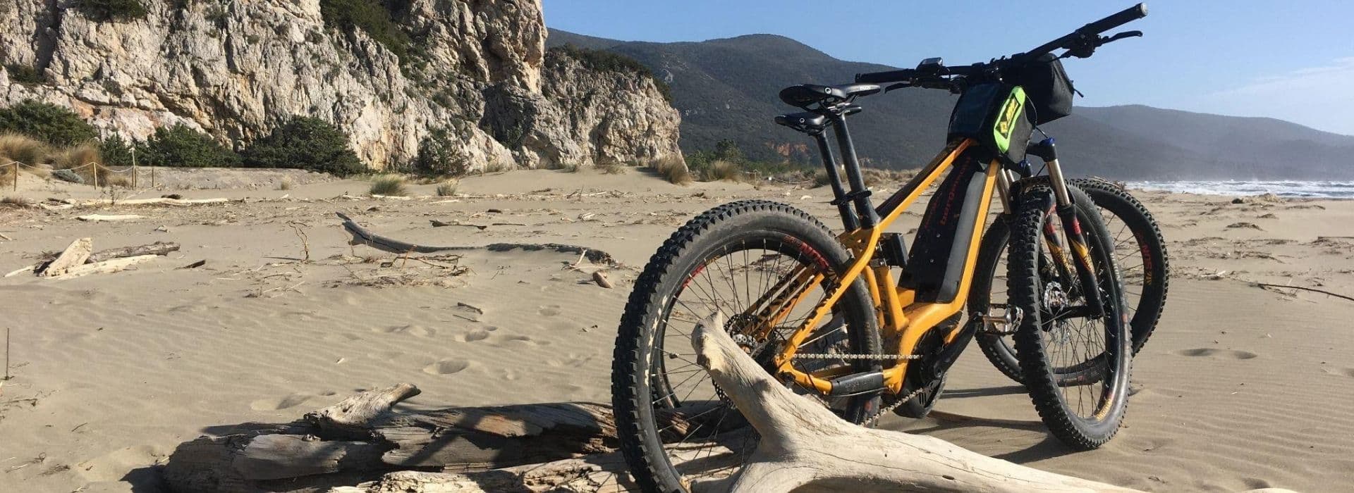 Due e-bike arancioni parcheggiate su un tronco di legno spiaggiato sulla spiaggia di Collelungo, nel Parco Naturale della Maremma, con falesie rocciose e colline boscose sullo sfondo