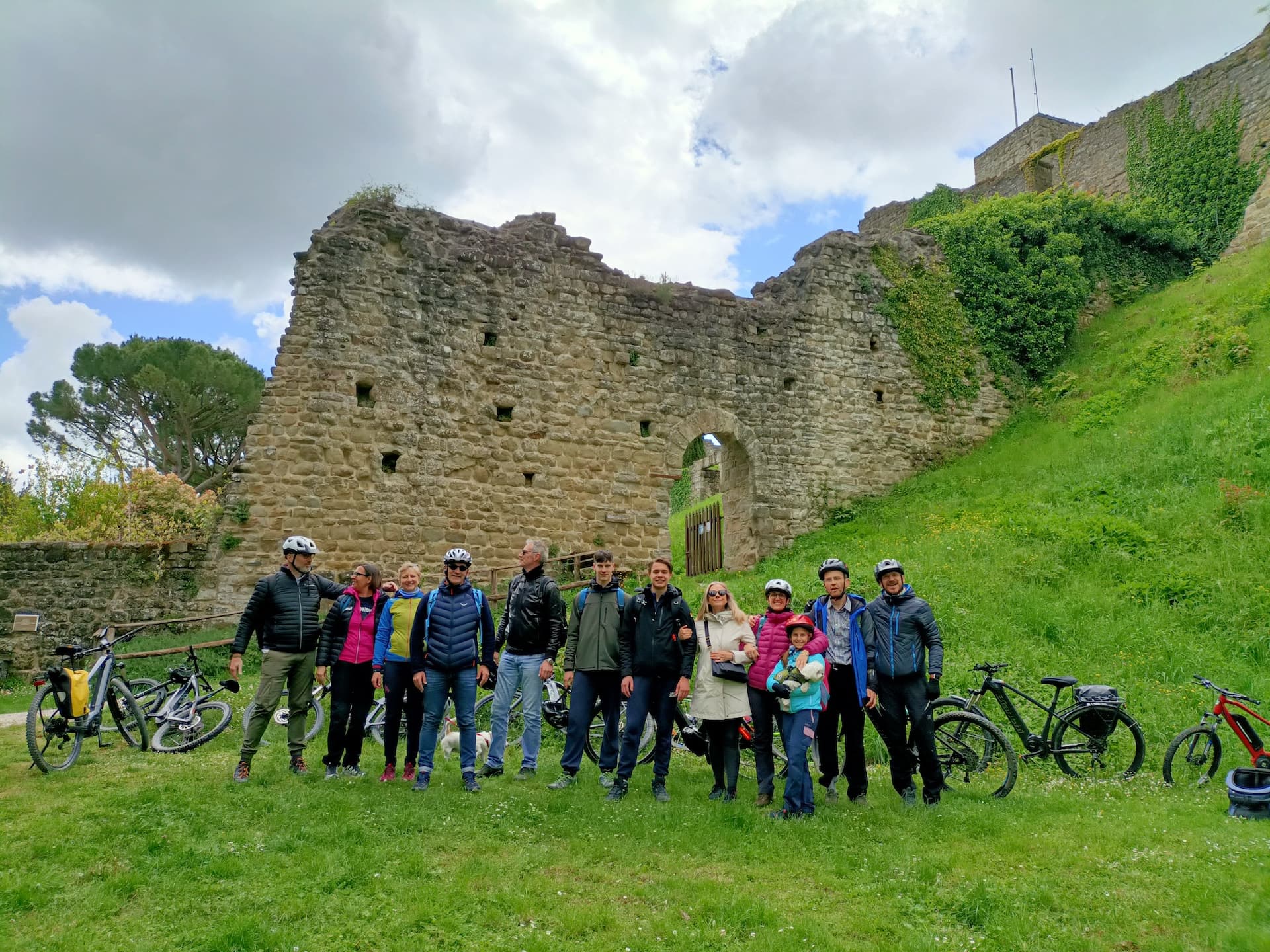 A group of eleven adults and a little girl wearing bike helmets pose on the grass in front of the ruined stone walls of Romena Castle, with e-bikes parked on either side, in Casentino