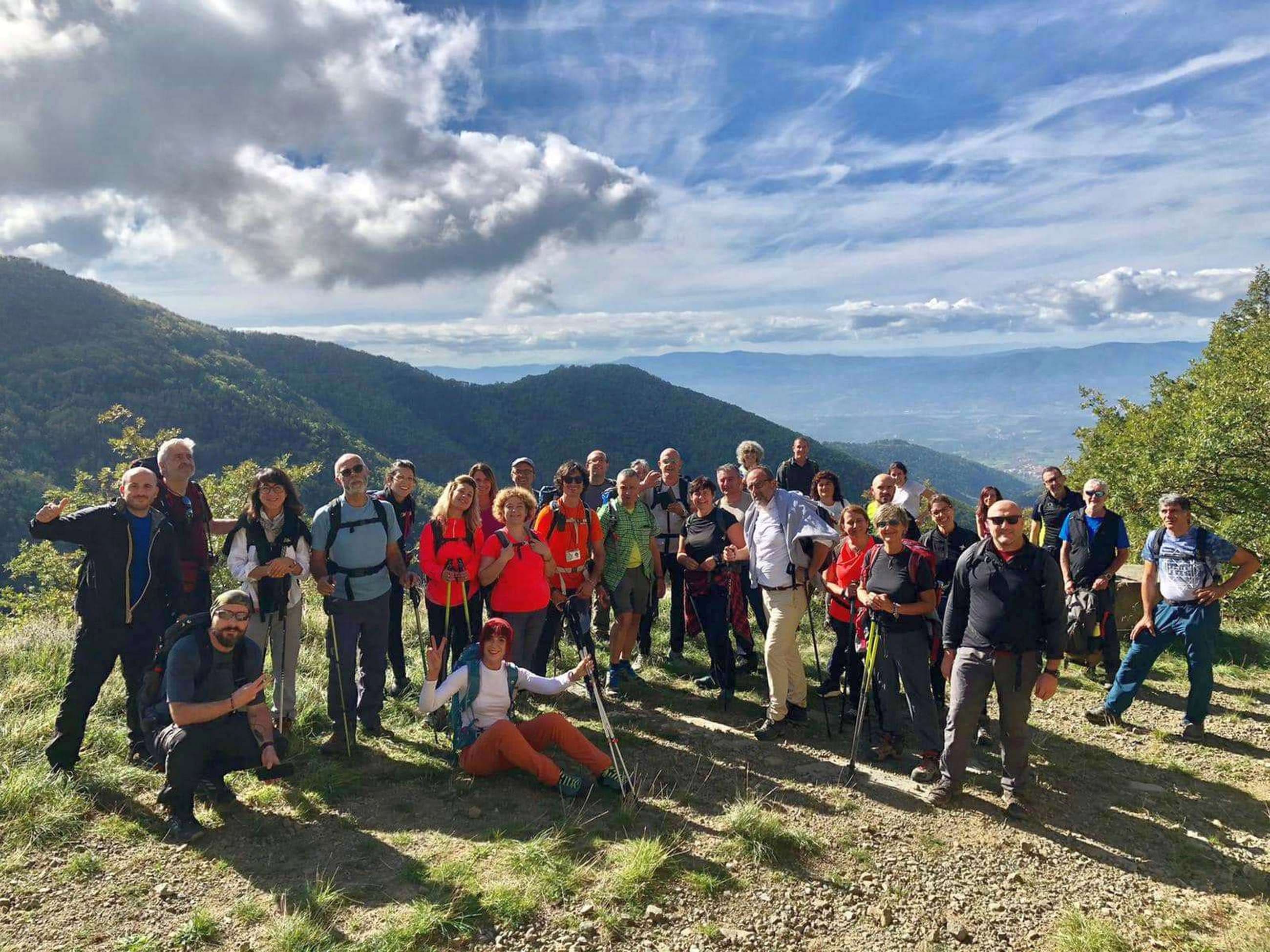 Gruppo numeroso di escursionisti in posa su un crinale aperto con bastoncini da trekking, con un ampio panorama di colline toscane boscose e cielo azzurro sullo sfondo