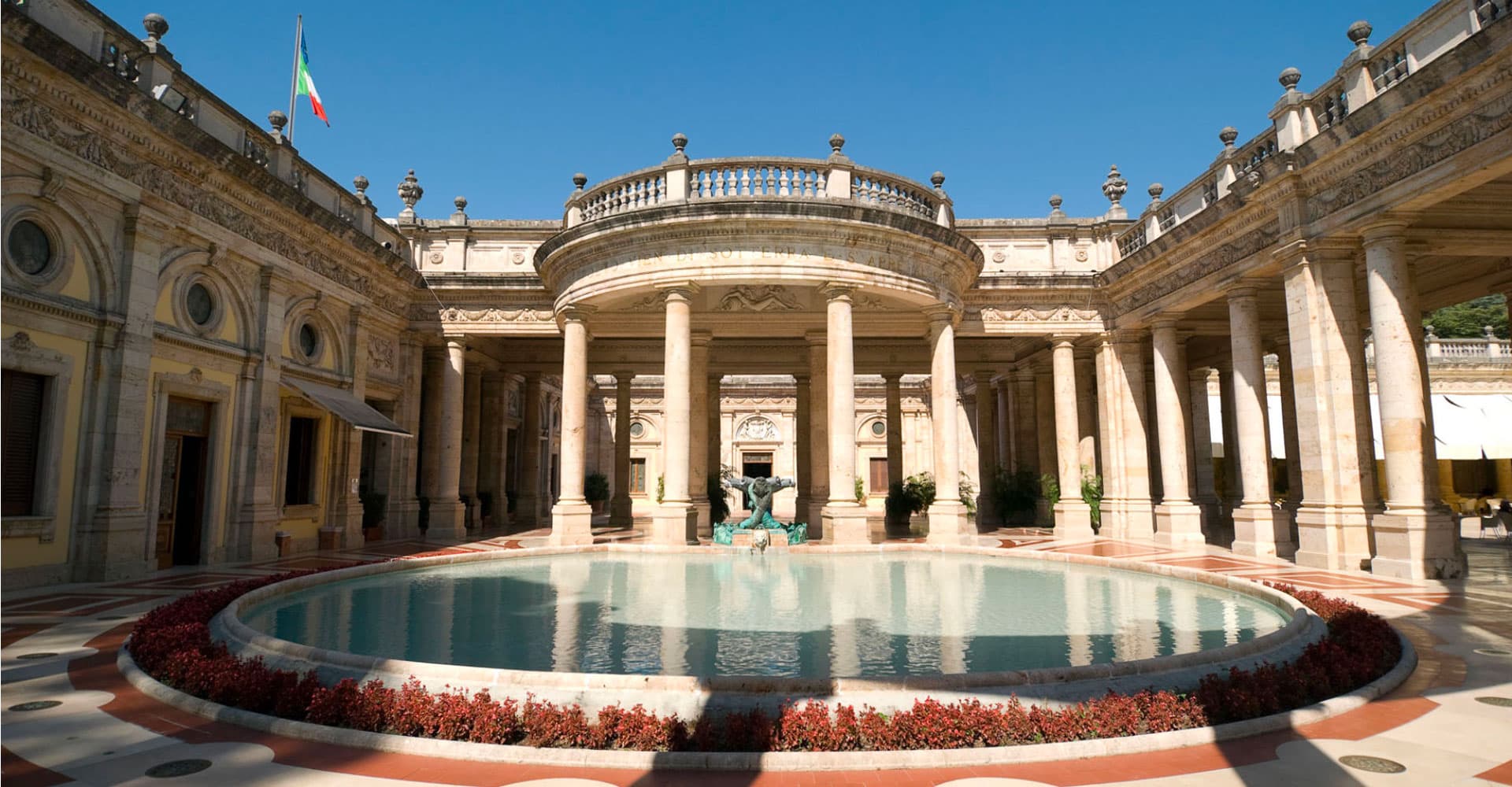 Cortile monumentale delle Terme Tettuccio di Montecatini Terme con vasca circolare centrale, fontana in bronzo, colonnato neoclassico e bandiera italiana, sede della Montecatini Gender Reveal Experience
