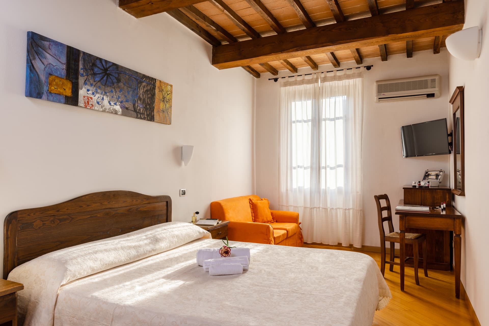 The image shows a cozy hotel bedroom that combines a rustic and classic style. The room features a ceiling with impressive exposed wooden beams, white walls, and a parquet floor. In the center, a double bed stands out with a dark wood headboard, a light-colored bedspread, and rolled towels, surmounted by a colorful, abstract rectangular painting. In the background, there is a window with white curtains, a vibrant orange small sofa in the corner, and a desk area on the right with a wooden chair, a mirror, a wall-mounted TV, and an air conditioner.