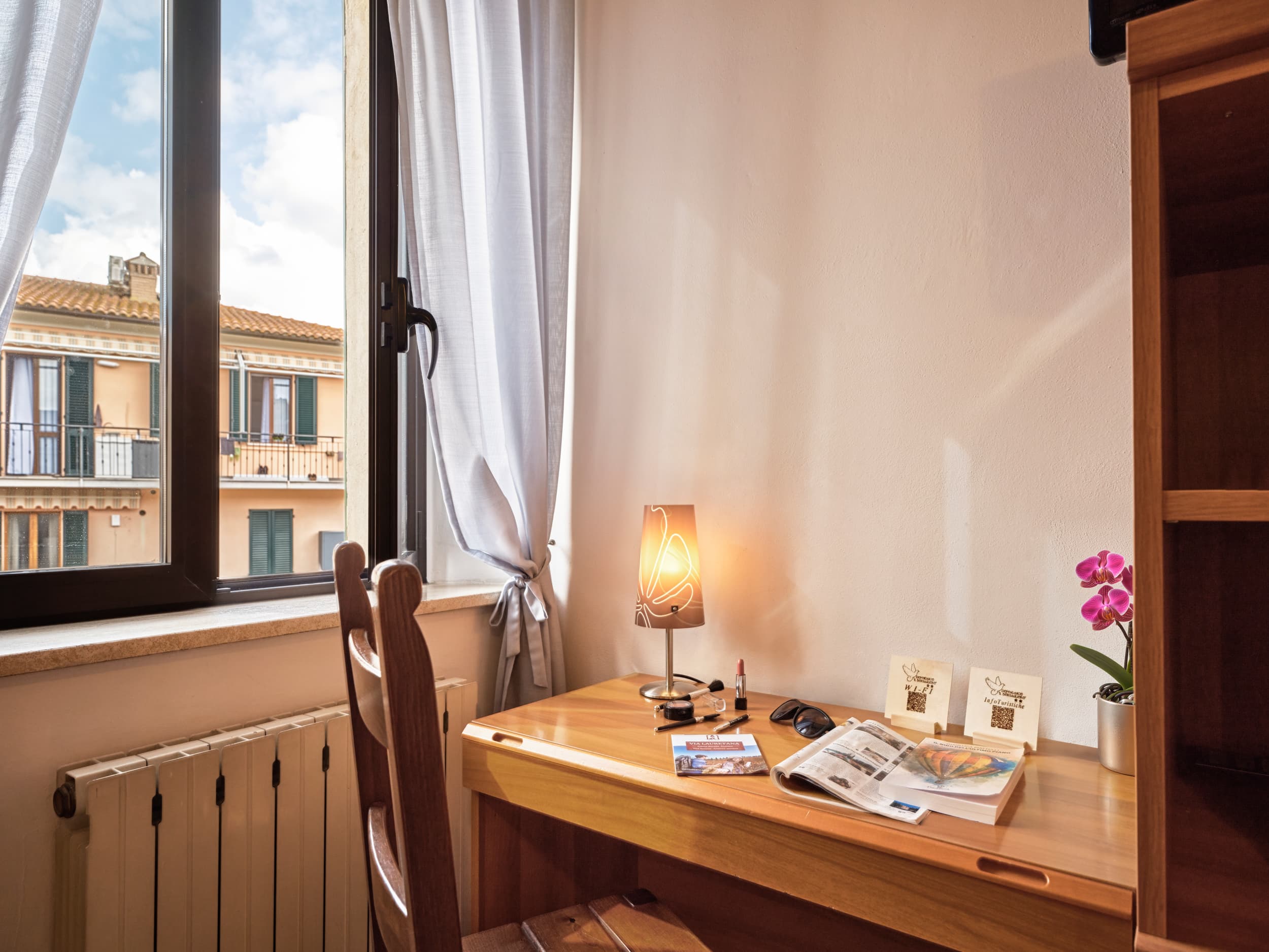 Angolo scrivania in legno di una camera d'albergo luminosa con sedia abbinata, lampada accesa, accessori, e vista su un borgo storico attraverso la finestra.