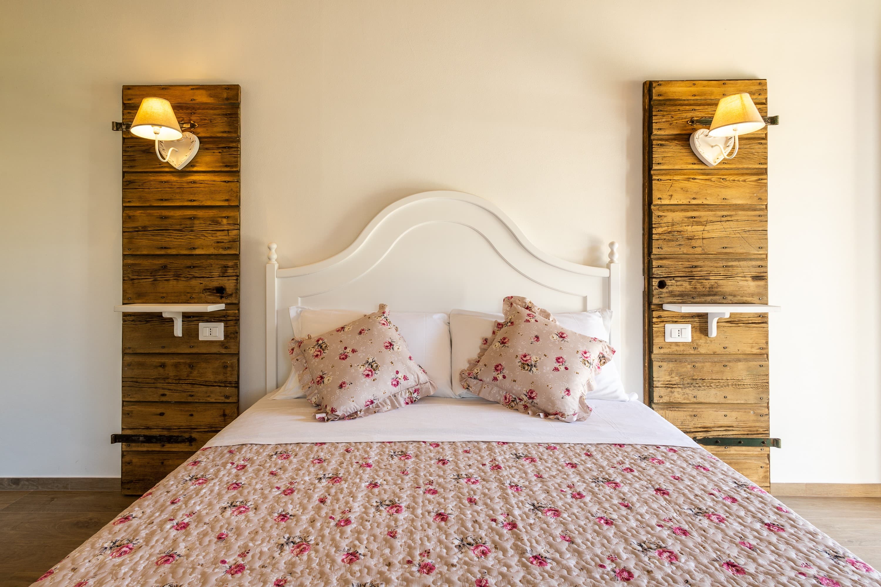 Frontal view of a country chic double bed featuring a shaped white wooden headboard. The bed is dressed in a quilted beige coverlet and matching pillows with a pink floral print and ruffle trim. On either side of the bed, two full-height vintage shutters made of dark reclaimed wood are mounted to the wall, serving as unique decorative backdrops for small white floating shelves and classic sconces with light fabric lampshades.