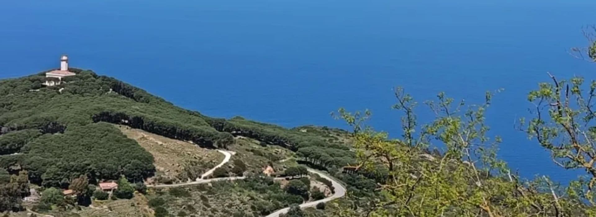 Strada sterrata che si snoda tra la macchia mediterranea verso il faro di Capel Rosso, sulla sommità di una collina dell'Isola del Giglio, con il mare azzurro dell'Arcipelago Toscano sullo sfondo
