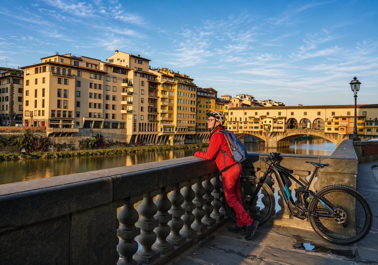 A cyclist with a backpack and helmet, wearing red gear, leans on the railing of a bridge in Florence, admiring the view of the Arno River, the historic buildings, and the famous Ponte Vecchio in the background, with her e-bike parked nearby