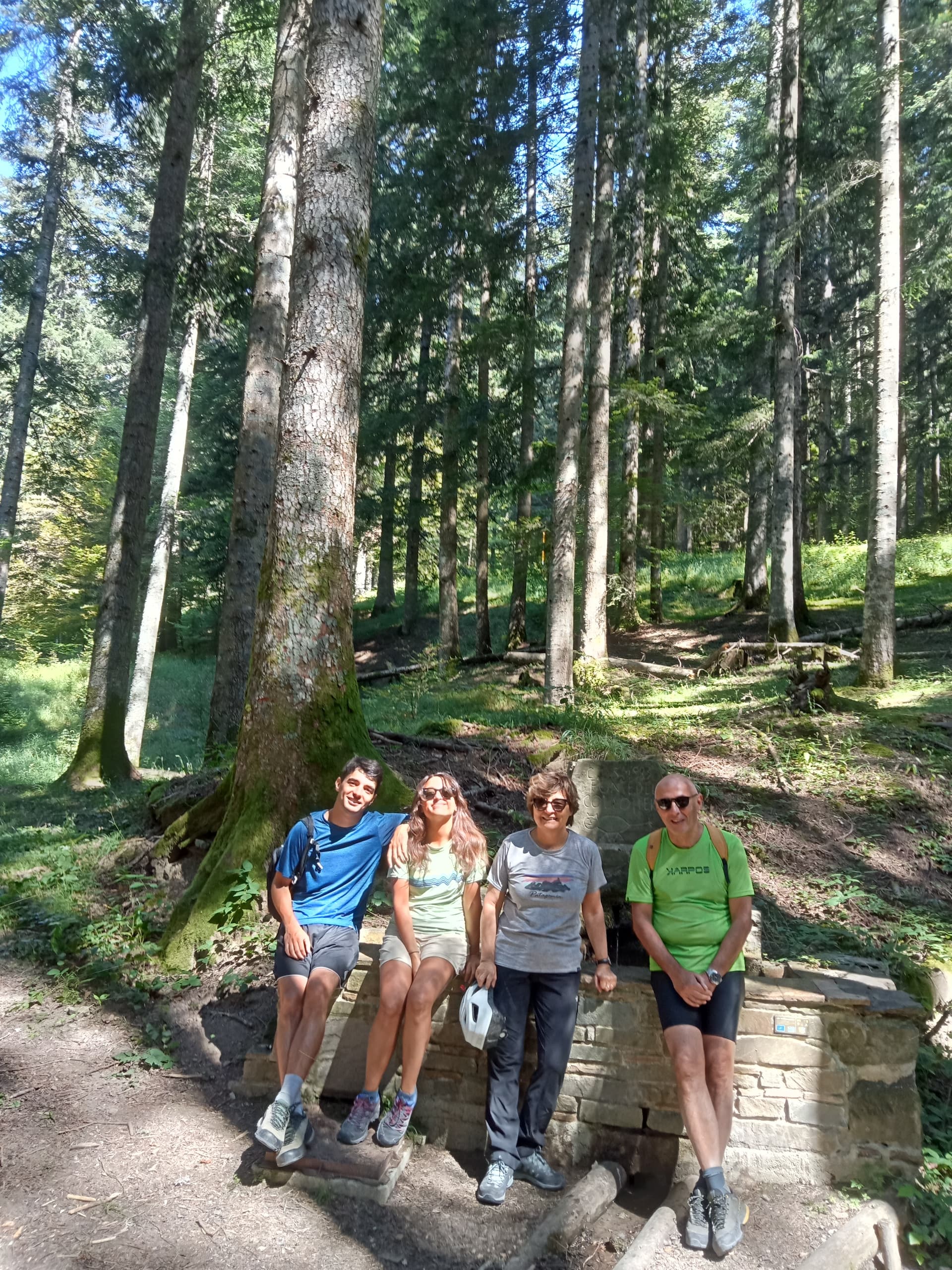 Quattro persone sedute sorridenti su una fontana in pietra all'interno di una fitta foresta di abeti bianchi del Parco Nazionale delle Foreste Casentinesi, in abbigliamento da escursione