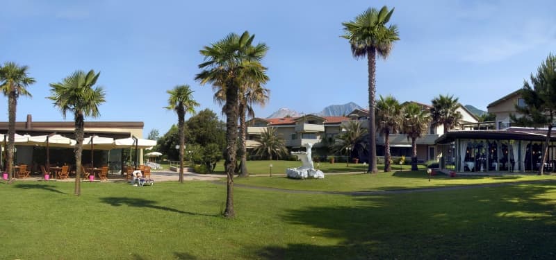 Giardino esterno di Hotel Villa Undulna con grandi palme, prato verde, tavoli all'aperto e montagne sullo sfondo