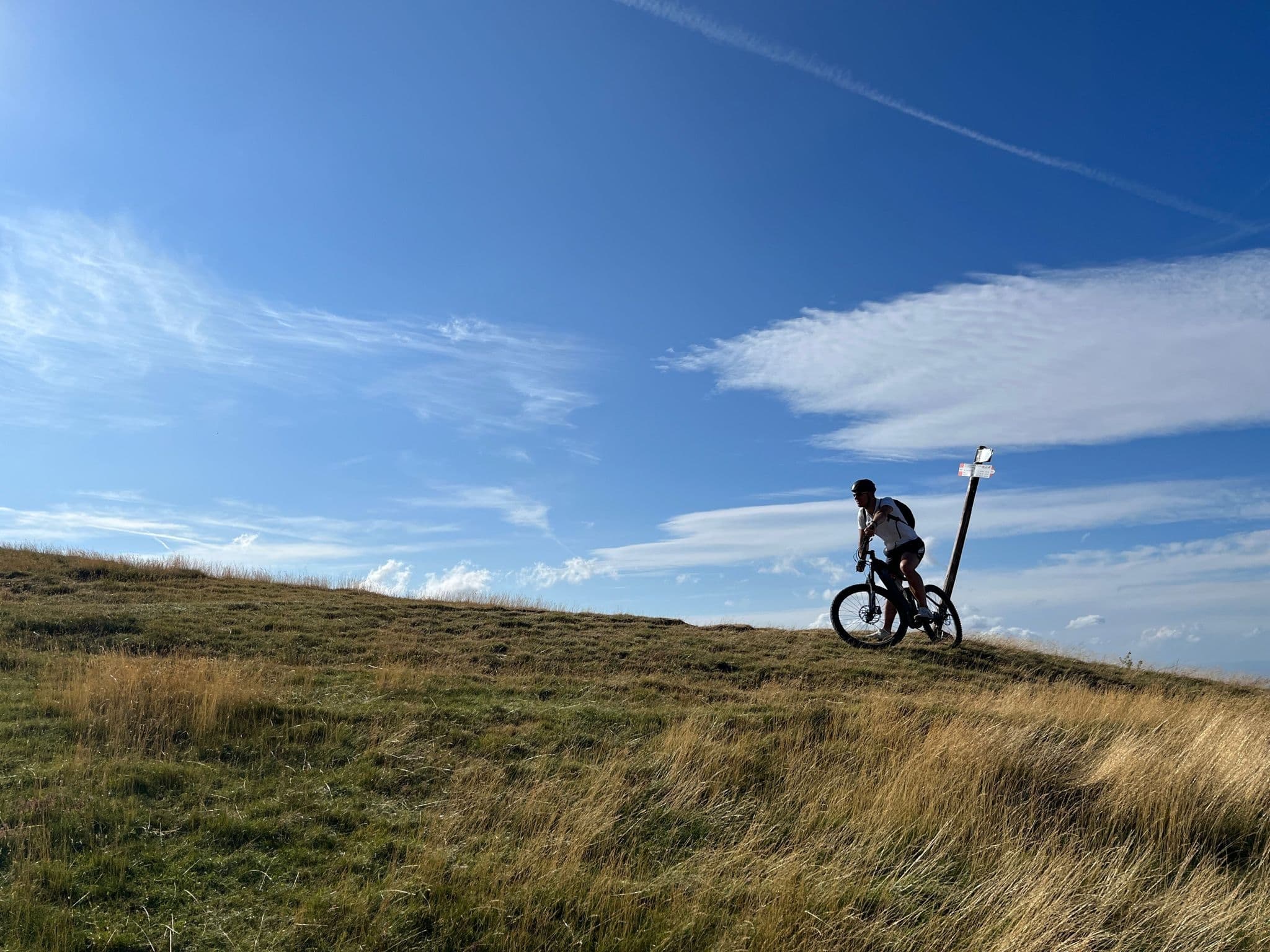 Cyclist in silhouette with e-bike atop a grassy ridge of Pratomagno next to a signpost, under a wide blue sky with clouds