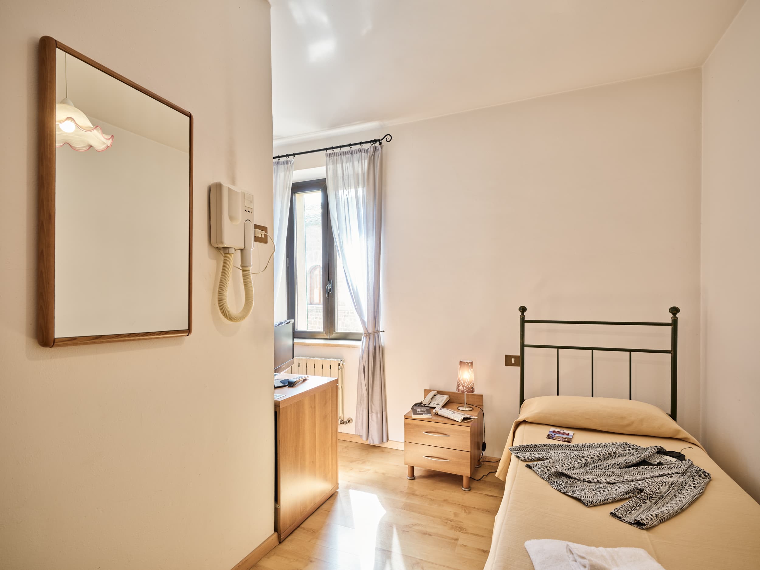 Camera singola di un hotel ad Asciano con letto in ferro battuto, comodino in legno, specchio a parete e finestra luminosa sullo sfondo.