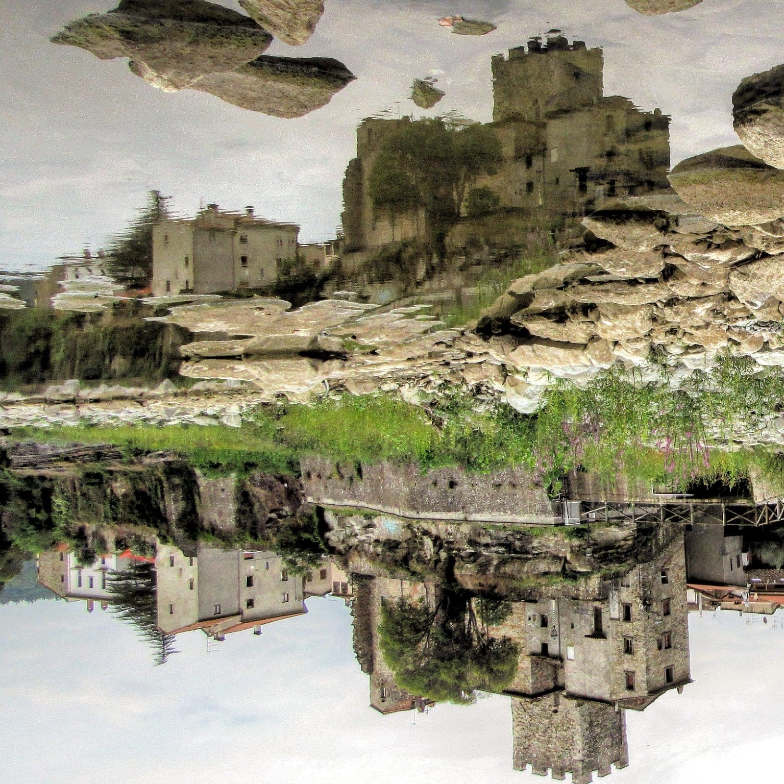 Riflesso capovolto nell'acqua del fiume Arno del Castello di Subbiano: la torre merlata, le costruzioni in pietra sul costone roccioso e la passeggiata lungo le mura appaiono speculari sulla superficie dell'acqua, creando un'immagine onirica e surreale