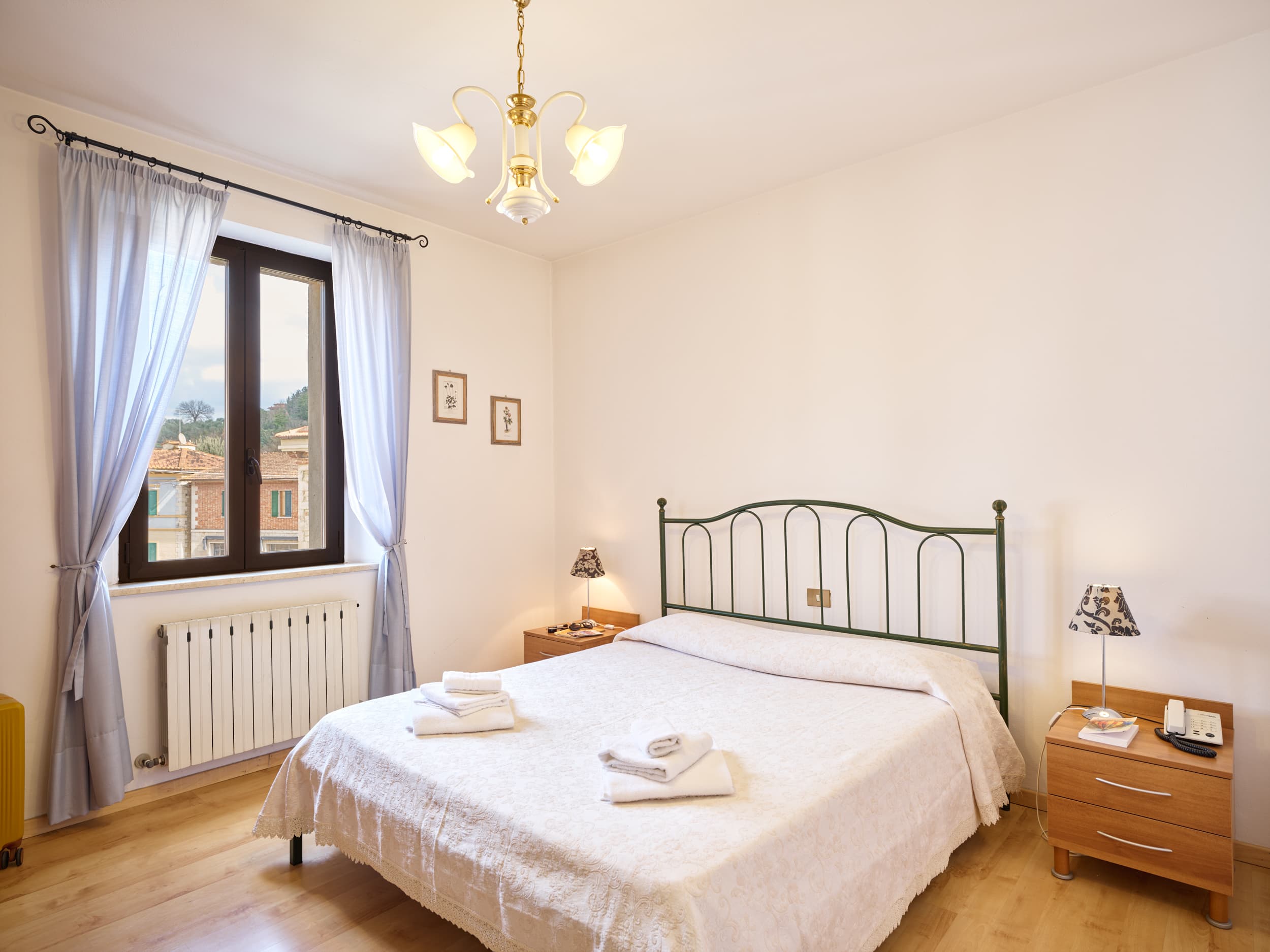 Camera da letto matrimoniale in stile toscano con letto in ferro battuto verde, pavimenti in parquet e vista panoramica su un borgo storico e vigne attraverso la finestra.
