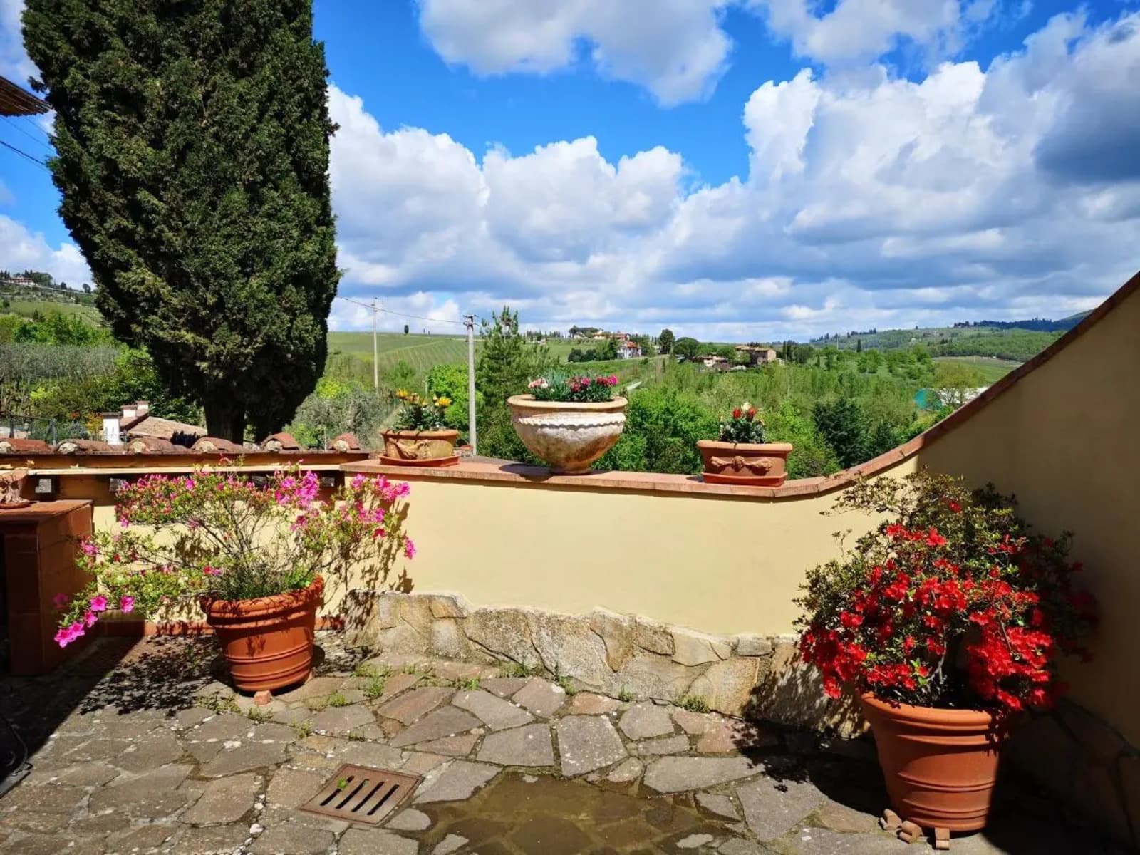 rustic stone terrace with paved flooring and an ochre-colored plastered retaining wall. The terrace is decorated with terracotta pots filled with blooming pink and red azaleas, along with classic planters aligned on top of the wall. A tall, iconic cypress tree stands on the left. The background features a sweeping panoramic view of the rolling green Chianti hills, dotted with vineyards, olive groves, and traditional farmhouses under a bright blue sky with large fluffy white clouds.