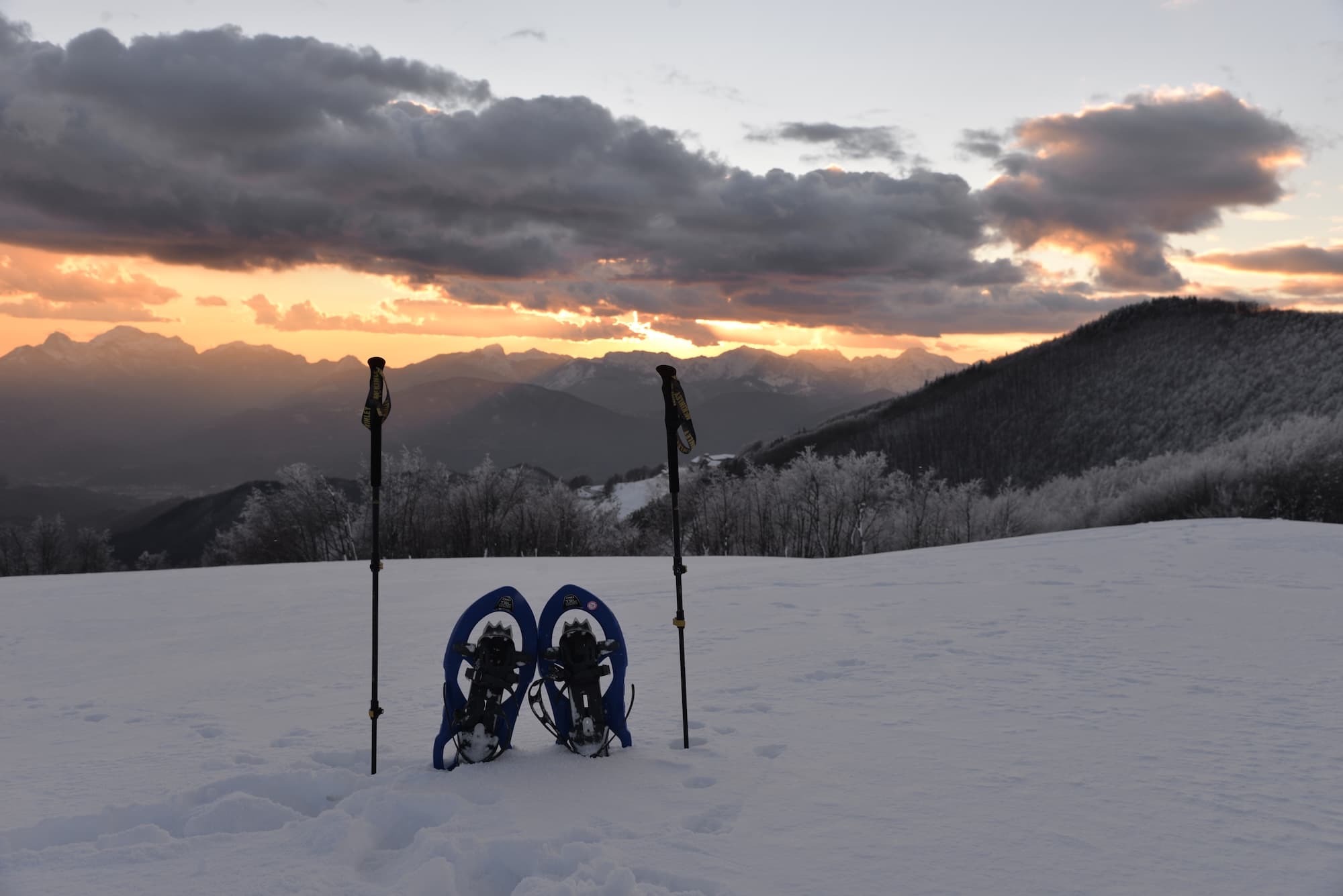 Ciaspole e bastoncini sulla neve al tramonto, con montagne innevate della Garfagnana e cielo nuvoloso sullo sfondo.